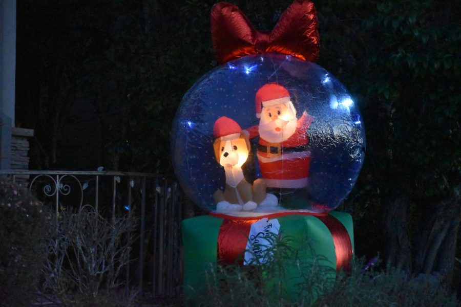 A blow-up Santa-in-a-snow-globe sits on a festive front lawn. Decorations like this remind everyone of the days when they believed in magic.