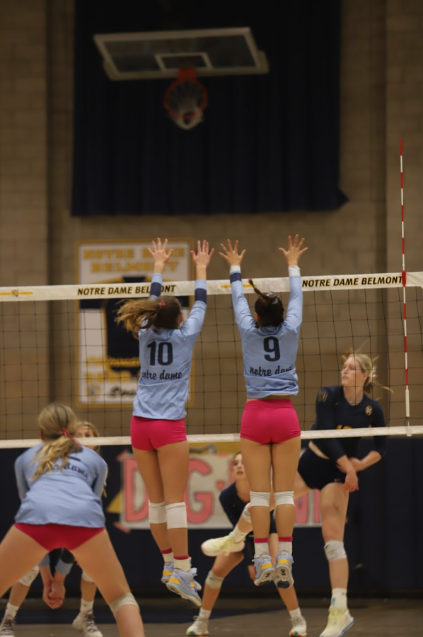 Varsity Volleyball players jump to block a hit during the Dig Pink game.