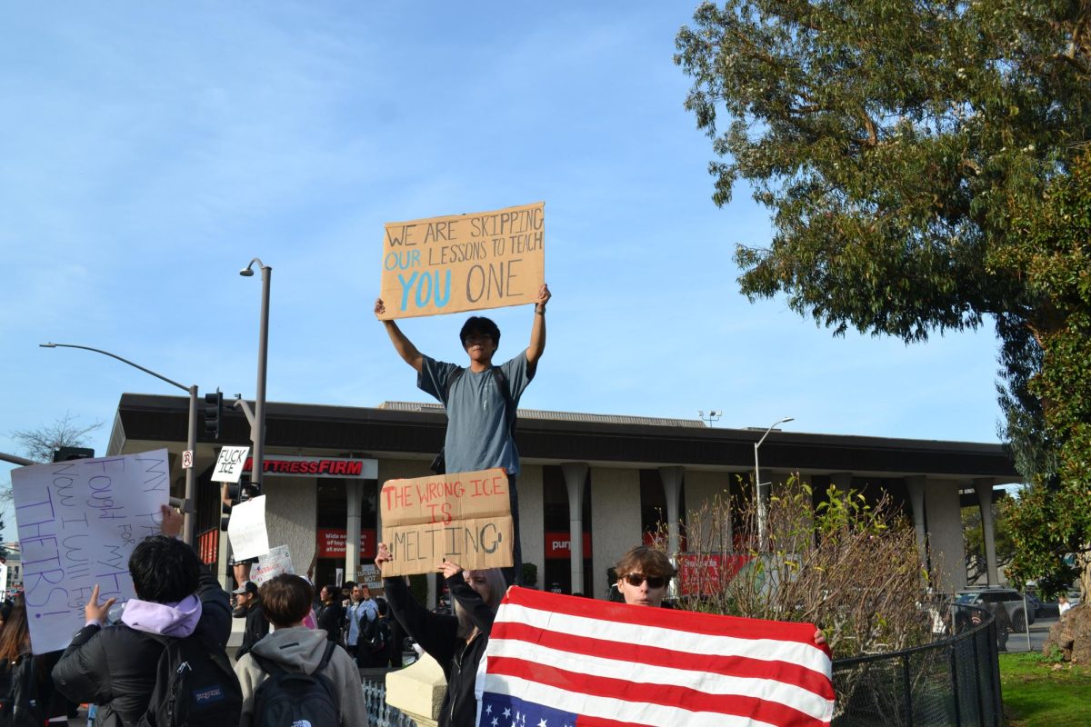A student holds a sign reading “We are skipping our lessons to teach you one” while standing above the crowd during a student-led protest over immigration enforcement policies in downtown San Mateo. 